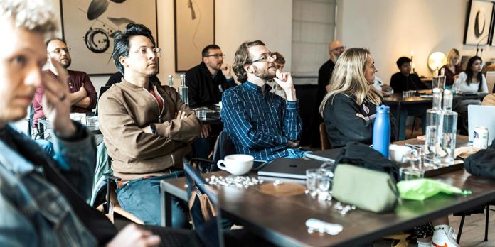 Grupo de personas adultas sentadas en una sala asistiendo a una clase o charla, con mesas, botellas de agua y portátiles, escuchando con atención.