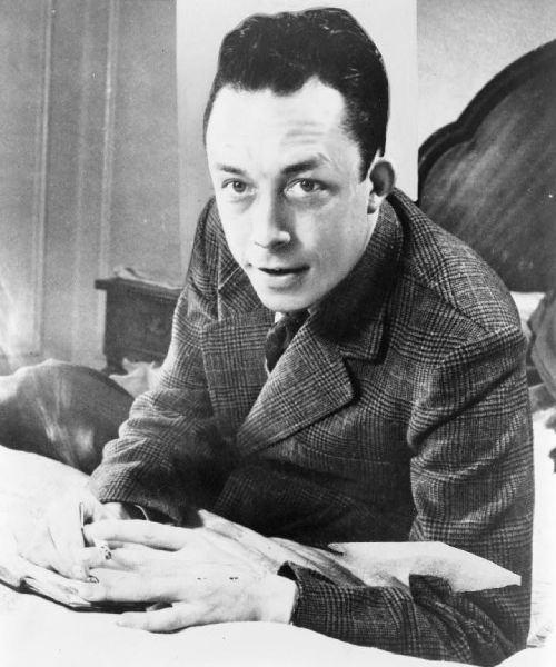 Albert Camus, sentado frente a un escritorio, mirando hacia la izquierda, fumando cigarrillo.