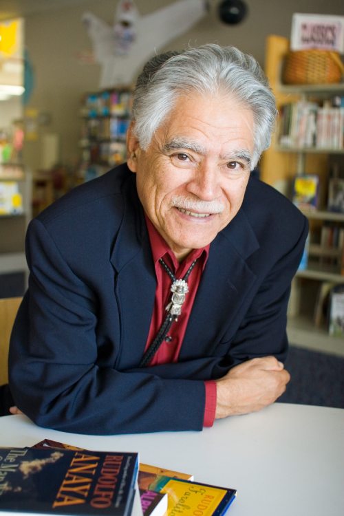 Rudolfo Anaya posa sus brazos sobre una mesa y sonríe a la cámara, al lado de sus libros.