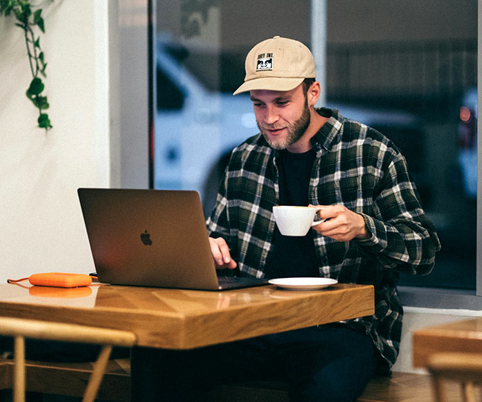 Hombre con gorra sentado en una cafetería frente a un ordenador portátil