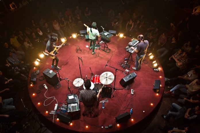 La imagen muestra a una banda tocando en un escenario circular rojo.