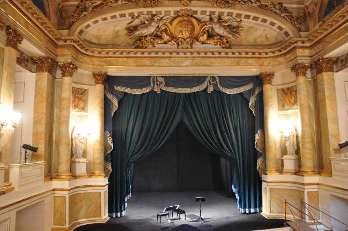 Fotografía de un teatro con escenario de proscenio, cortinas azules y barrotes dorados con blanco.